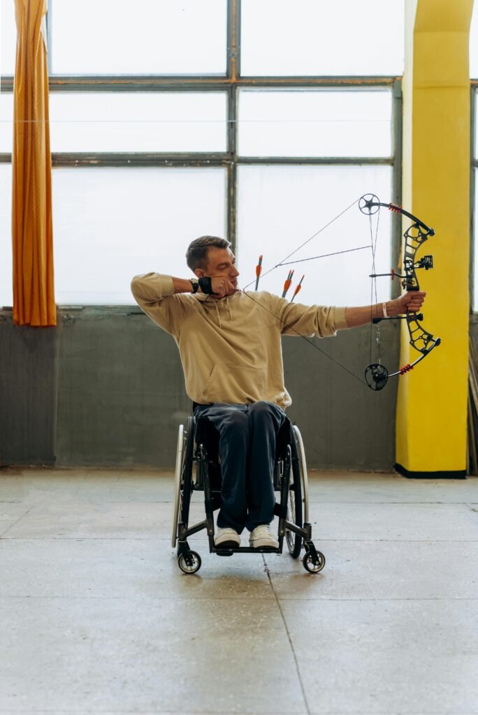 Man in a wheelchair practicing archery indoors with a compound bow.